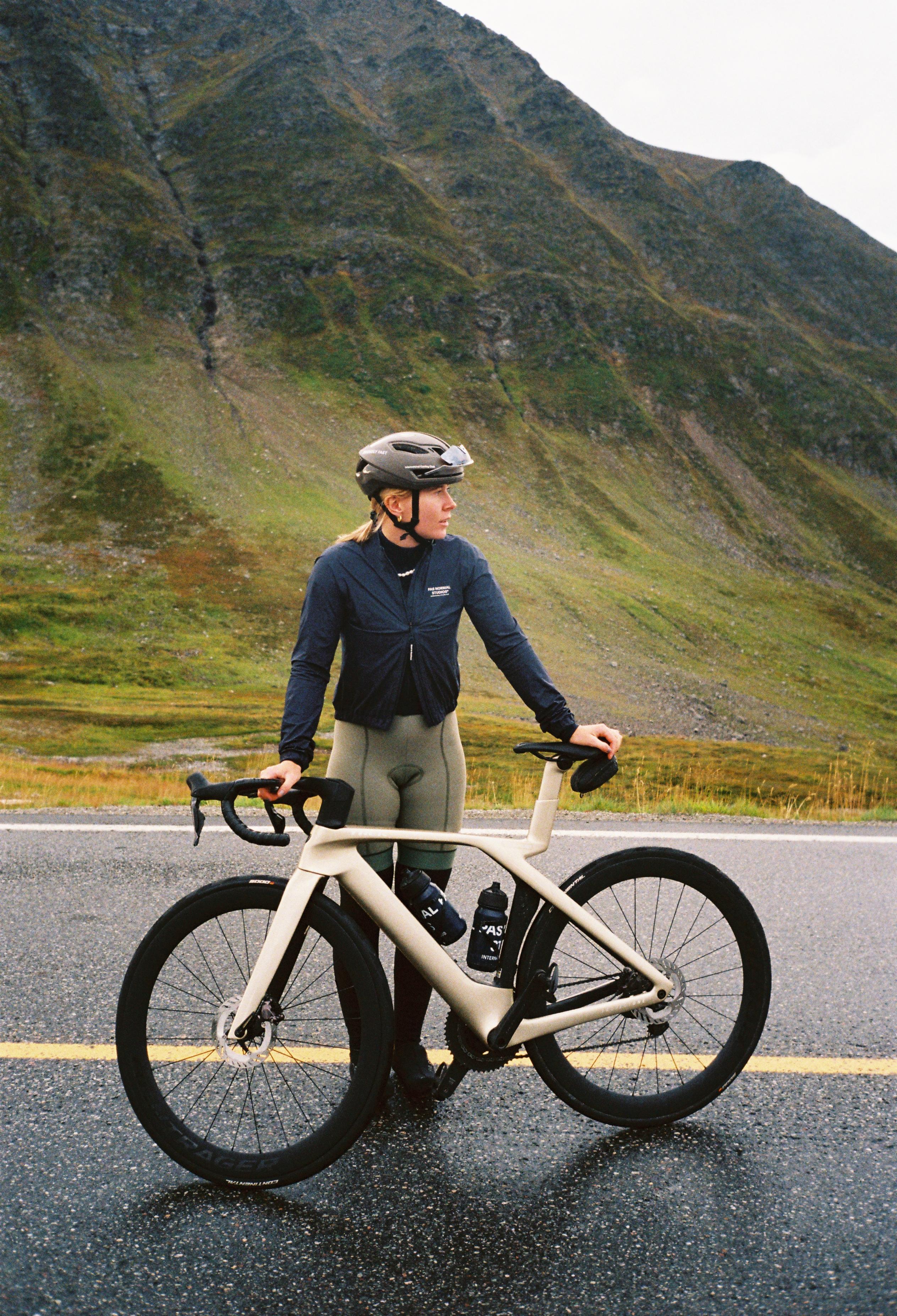 Model wearing navy cycling jacket with grey leggings standing with bike on mountain road