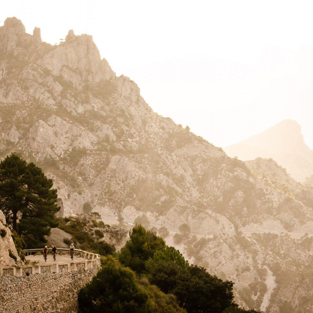 Three cyclists on mountain road with dramatic rocky peaks in golden sunset light