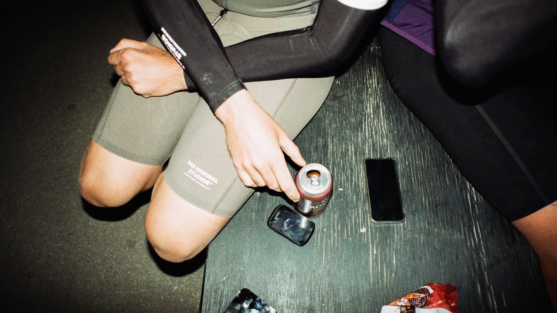Model wearing olive green cycling shorts with leg branding, seated at wooden table with coffee cup and phone
