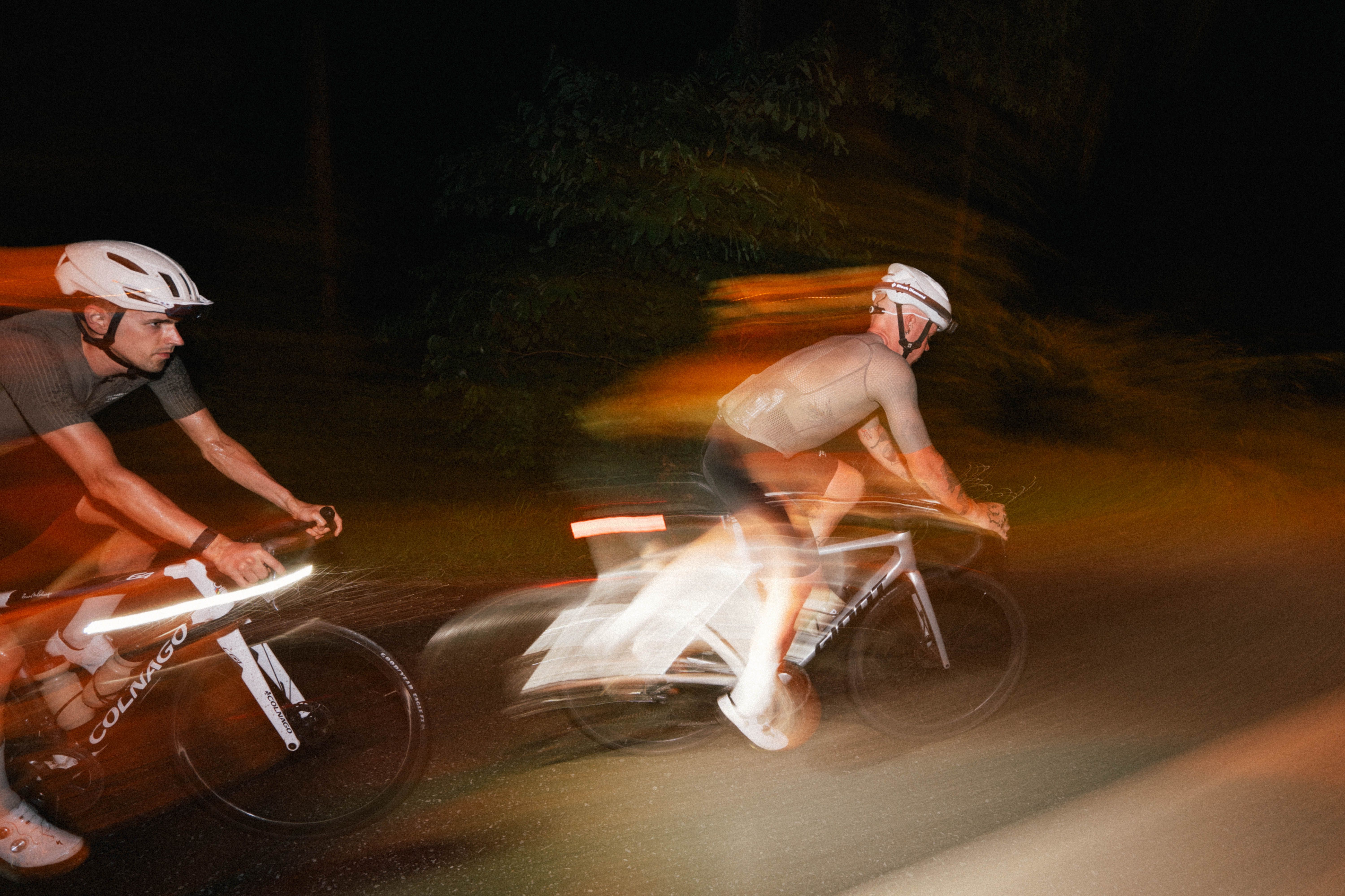 Two cyclists racing on time trial bikes at night with motion blur and orange lighting effects