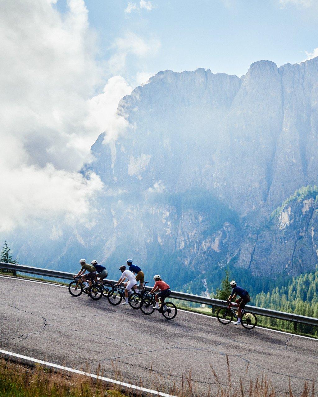Group of cyclists in colorful jerseys riding on mountain road with dramatic alpine peaks and clouds