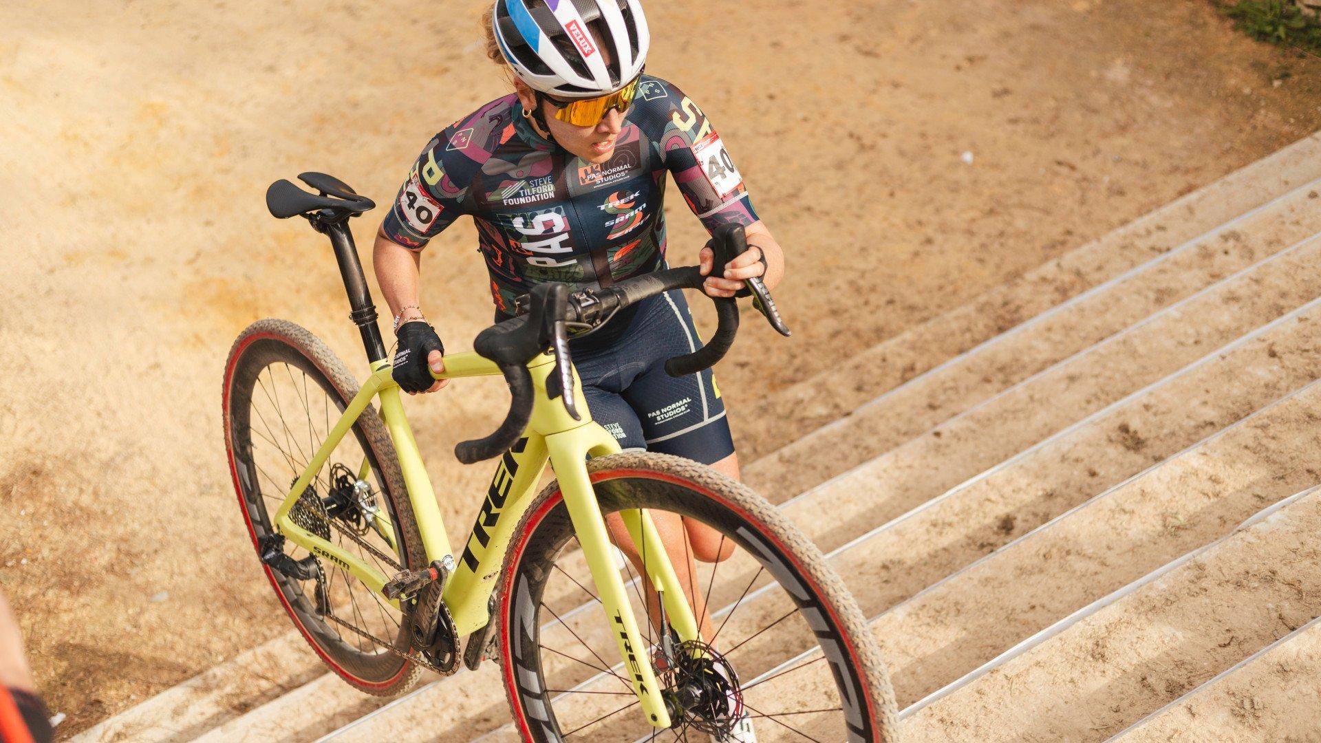 Model wearing colorful geometric print cycling jersey and navy shorts during cyclocross race on sandy terrain