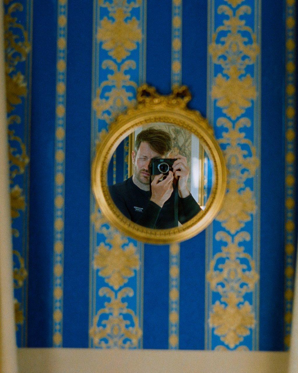 Model taking photo with camera, reflected in ornate gold mirror against blue patterned wallpaper