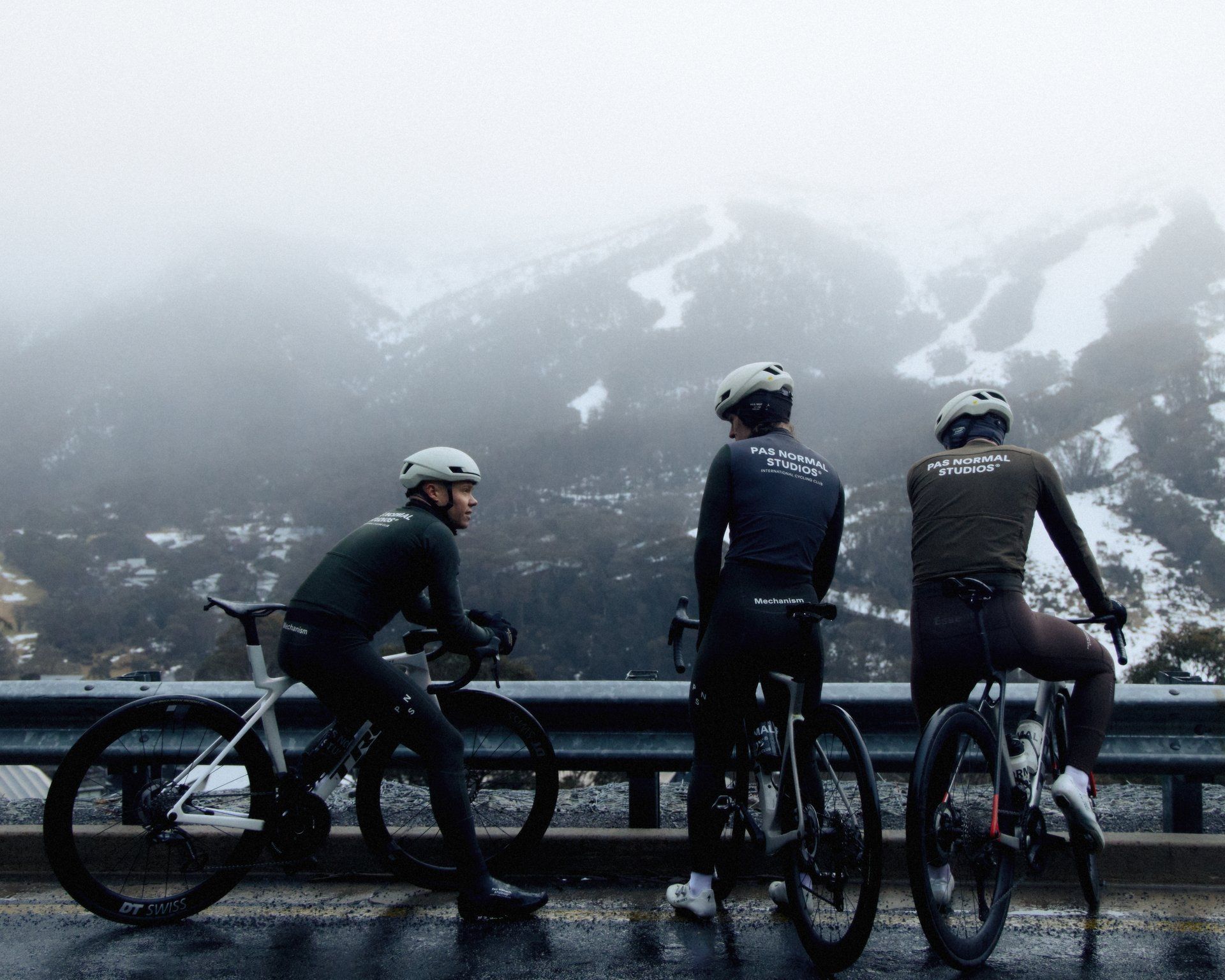 Three cyclists wearing dark winter cycling gear with back branding, standing with bikes against snowy mountain backdrop