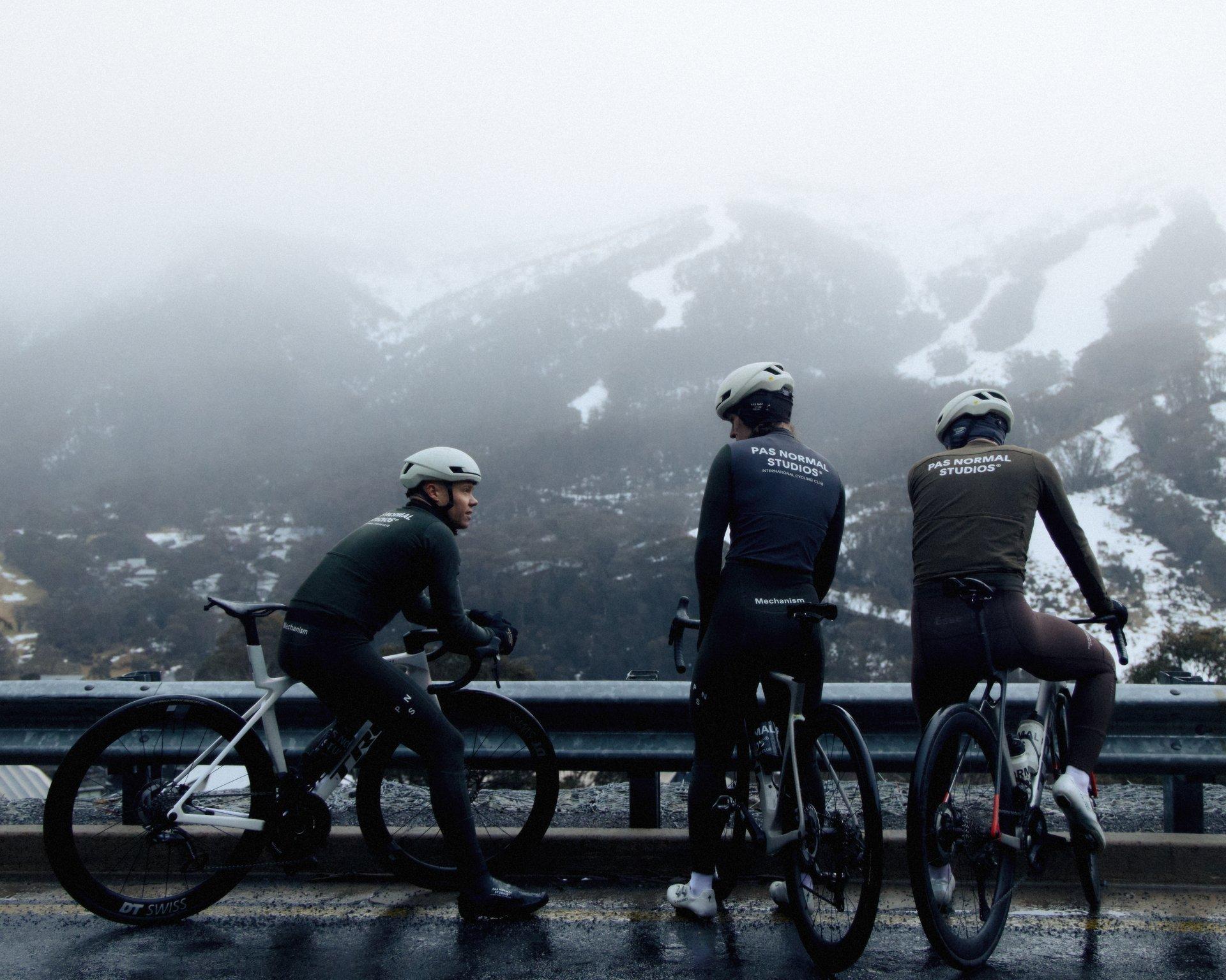 Three cyclists wearing dark winter cycling gear with back branding, standing with bikes against snowy mountain backdrop