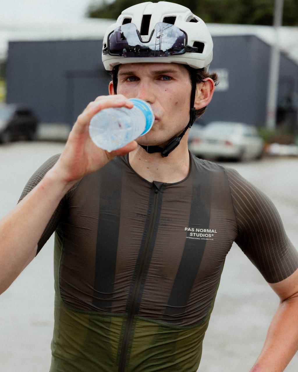 Model wearing dark green textured cycling jersey with branding, drinking from water bottle while wearing white helmet