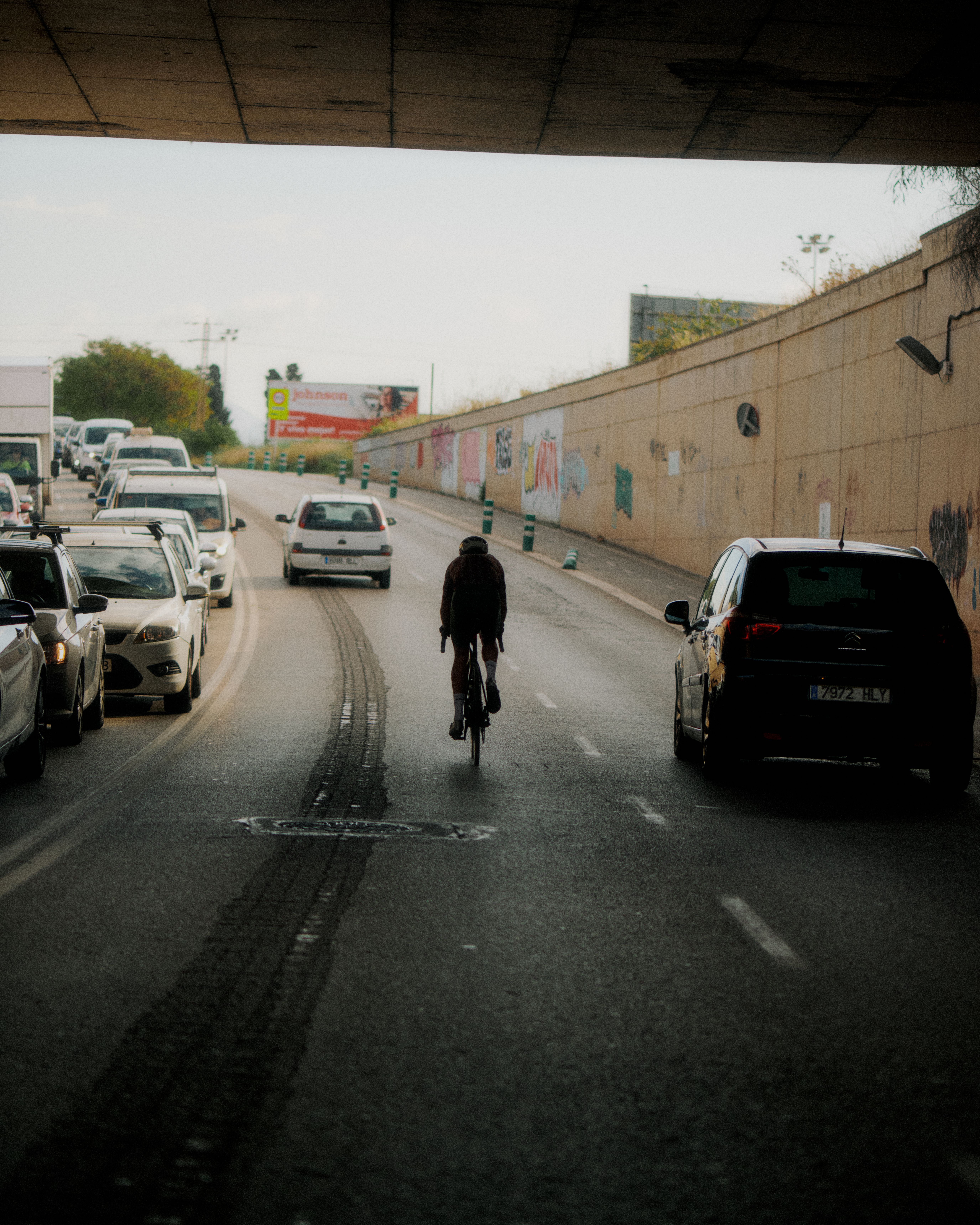 Cyclist in dark clothing riding through urban underpass with traffic and graffitied walls