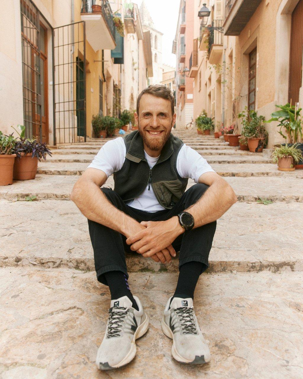 Model wearing dark green vest over white t-shirt with black shorts, sitting on stone steps in Mediterranean village