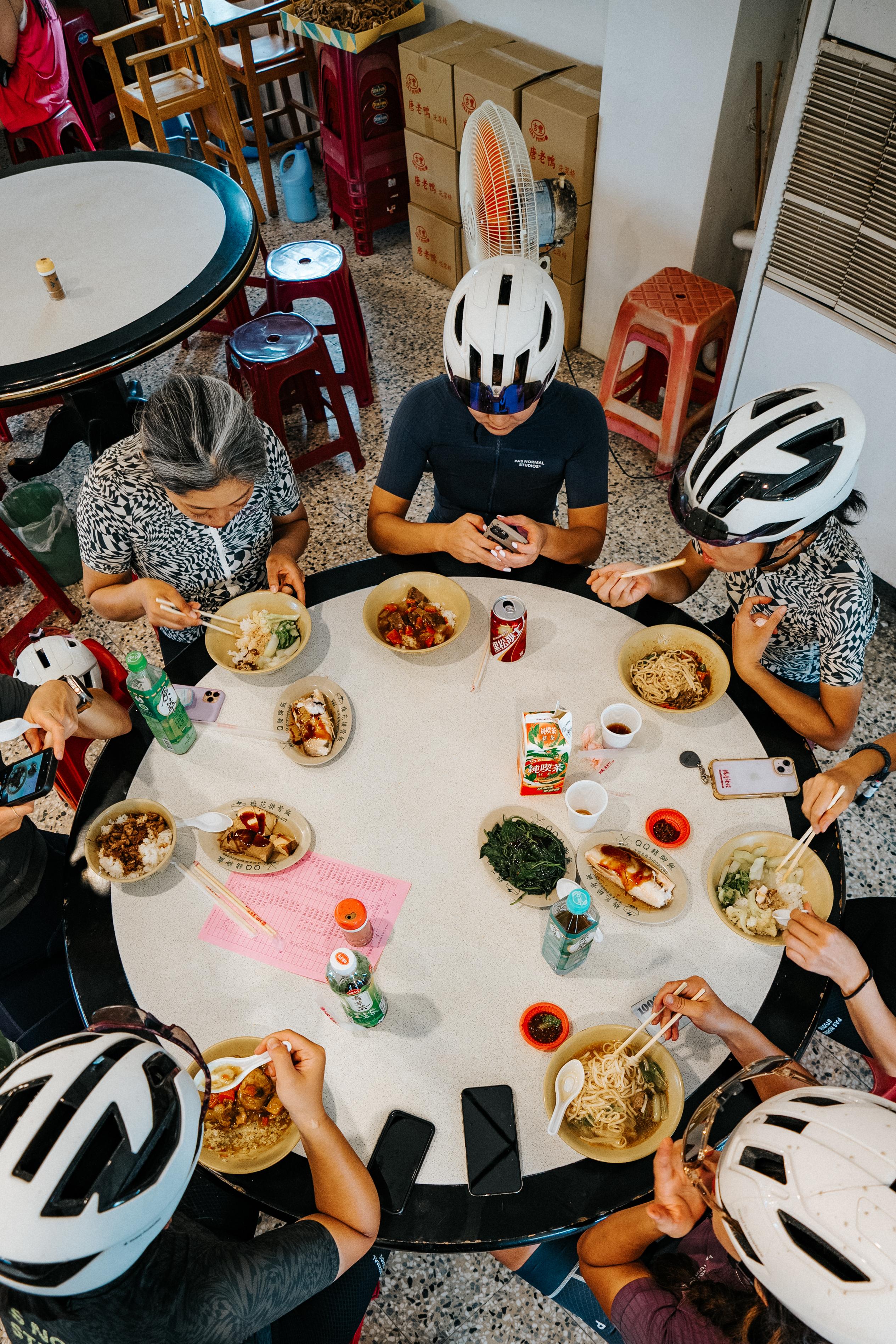 Cyclists in helmets and cycling apparel sharing a meal around table during group ride stop