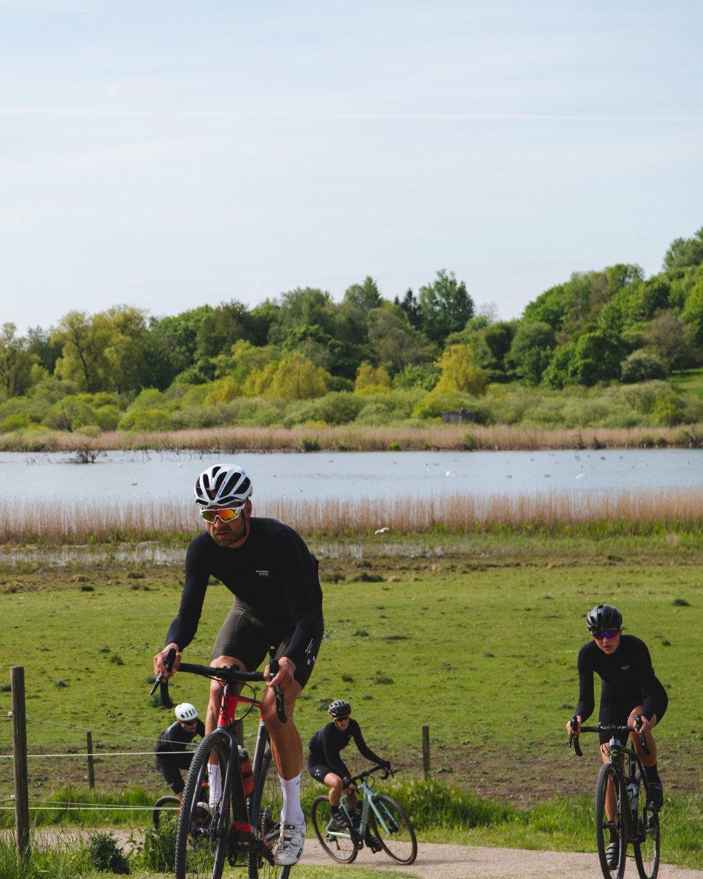 Cyclists wearing black long sleeve jerseys riding along lakeside path in countryside setting