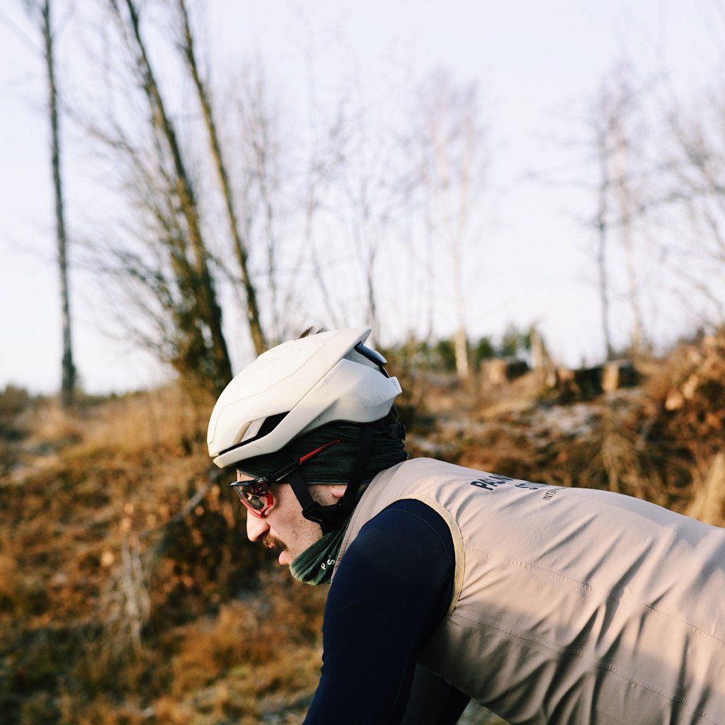 Model wearing white cycling helmet and grey vest with navy base layers in aerodynamic position outdoors