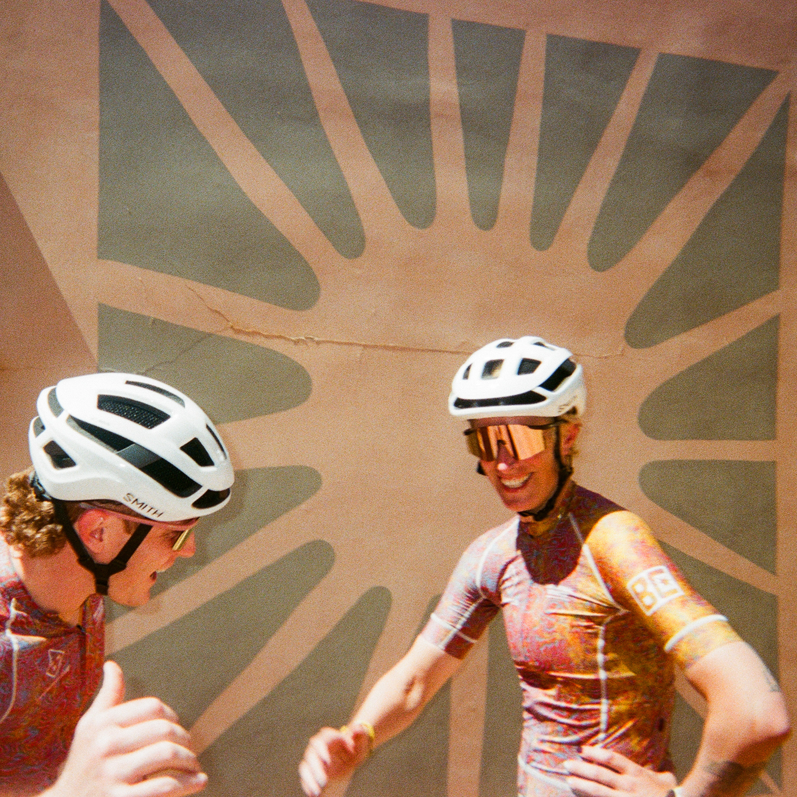 Two cyclists in white helmets against desert-colored wall with palm shadow pattern