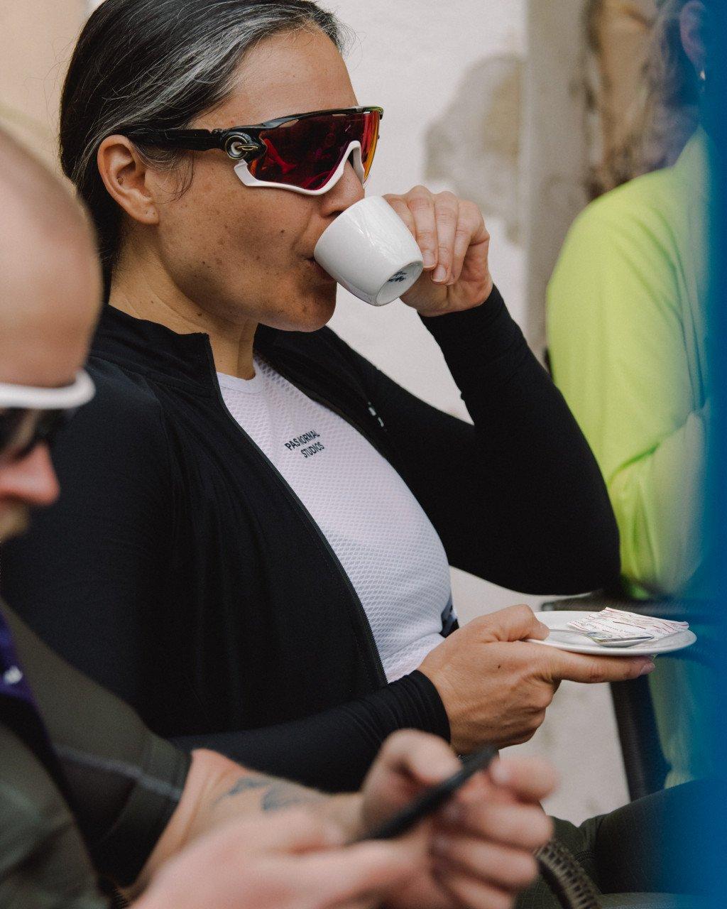 Model wearing black cycling jacket over white base layer drinking coffee