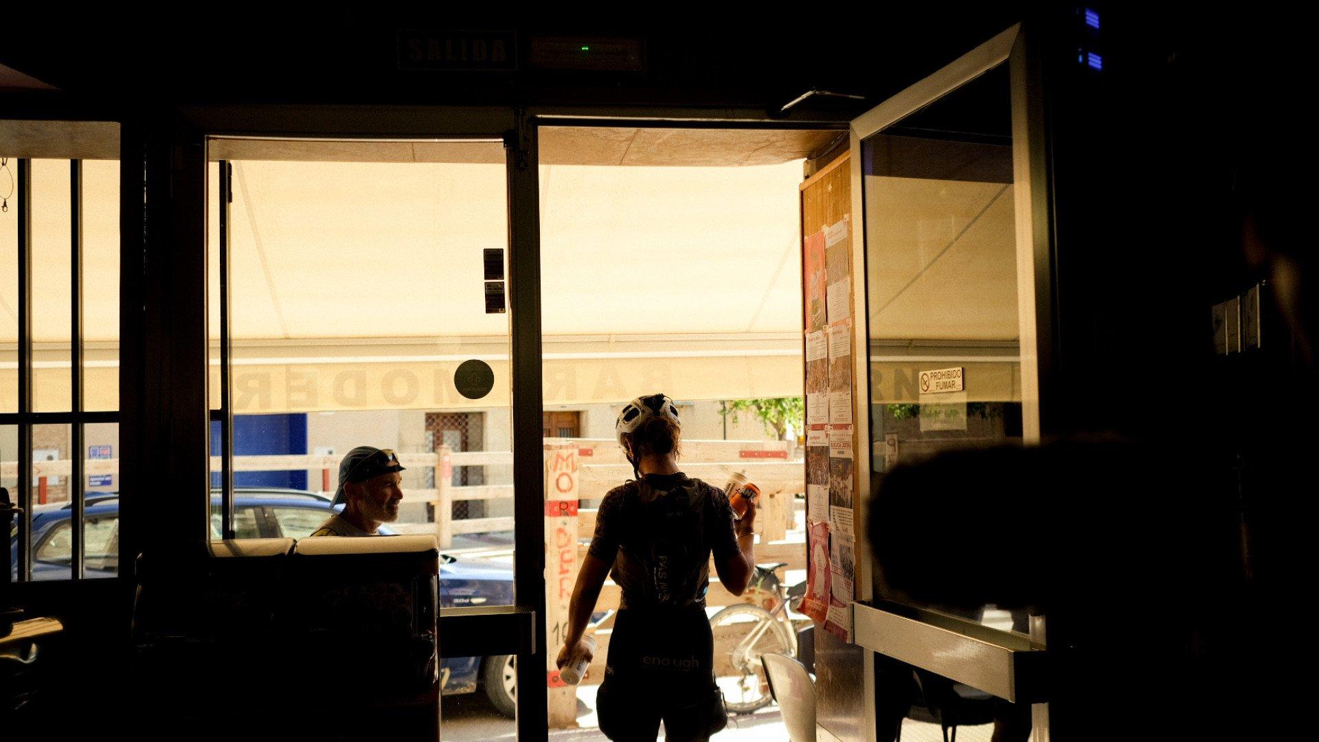 Model wearing dark cycling apparel in coffee shop doorway, sunglasses on head