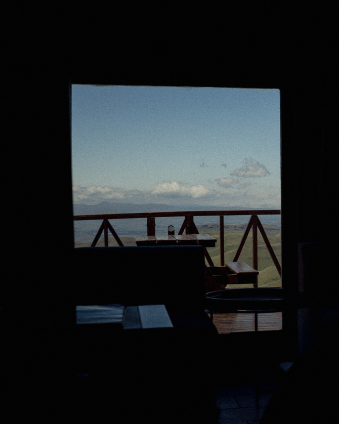Interior view through window showing wooden deck overlooking mountainous landscape at dusk