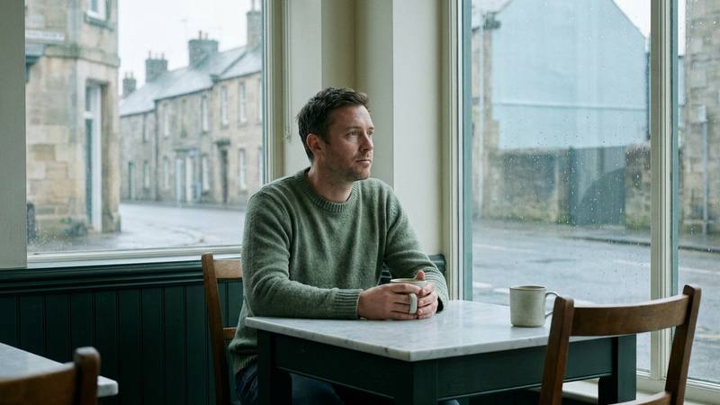 A man sitting alone at a cafe table by a window, looking away, an untouched second mug across from him