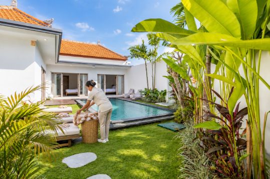 Housekeeper carrying fresh towels across the lawn beside a private pool in a Bali villa