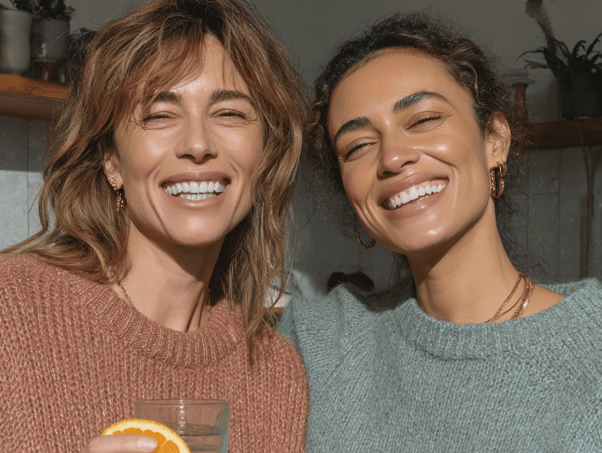 Two women laughing together in a kitchen, one holding a glass with an orange slice