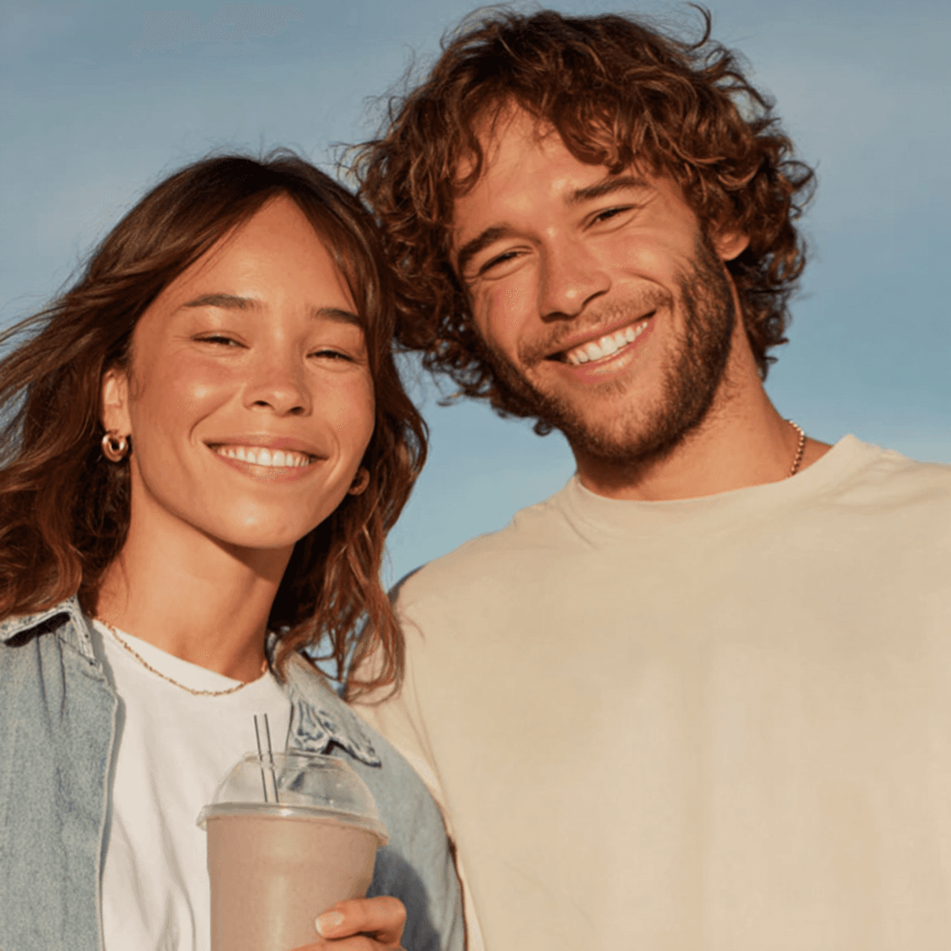 A young couple smiling outdoors against a blue sky, one holding a drink
