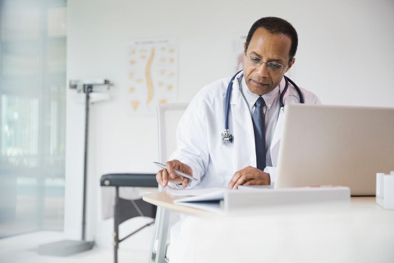 Male doctor wearing a stethoscope reviewing his notes with a laptop open in the foreground