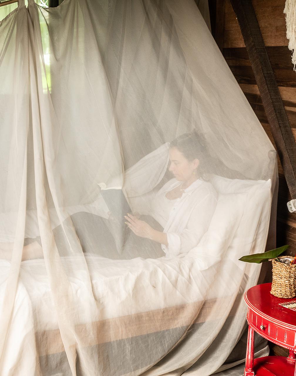 A woman sat in bed in a cabin reading a book under a mosquito net