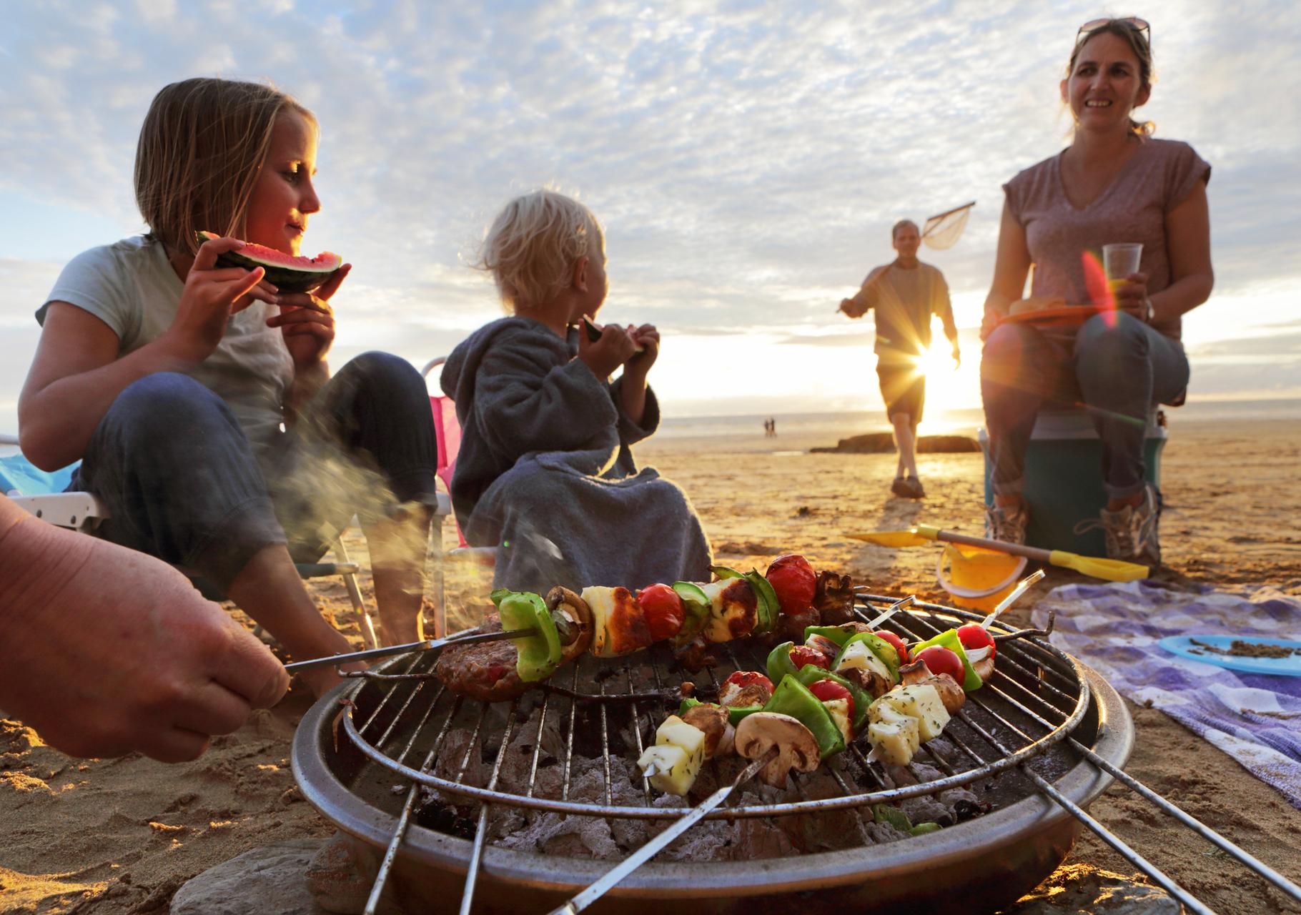 Young child sat next to a woman eating by a bbq on the beach, a man is walking towards them with a net