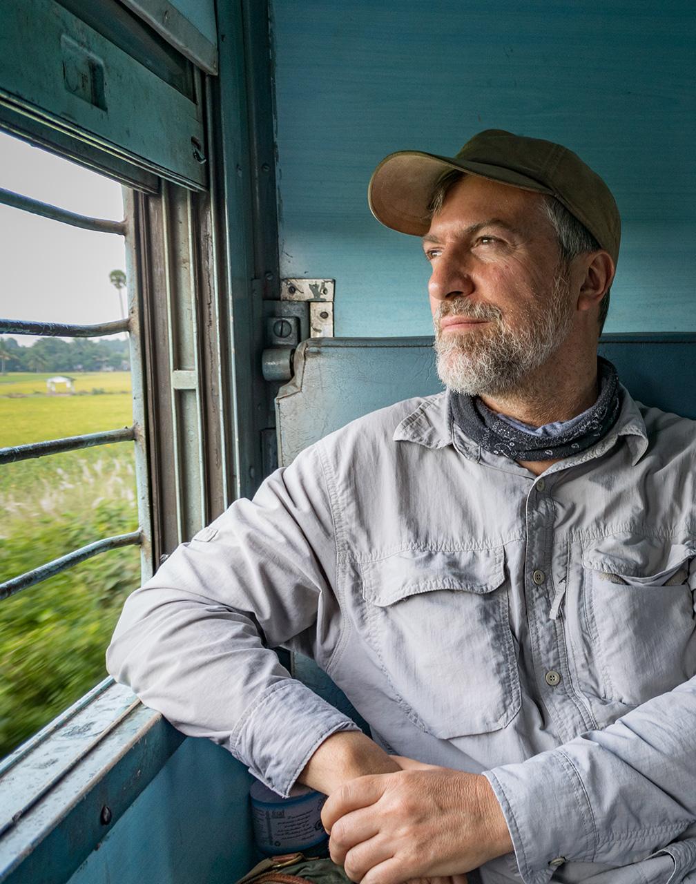 A man sat in the back of an old vehicle with his backpack next to him looking out a window with bars across