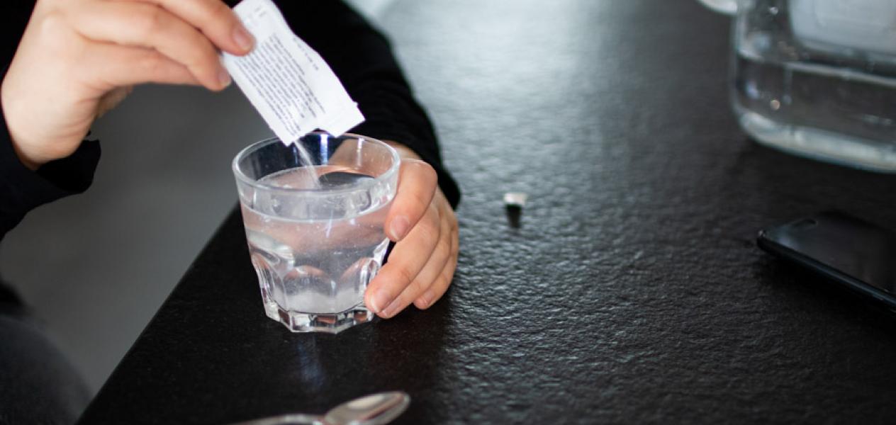 A hand tipping a sachet of powder into a glass of water on a counter