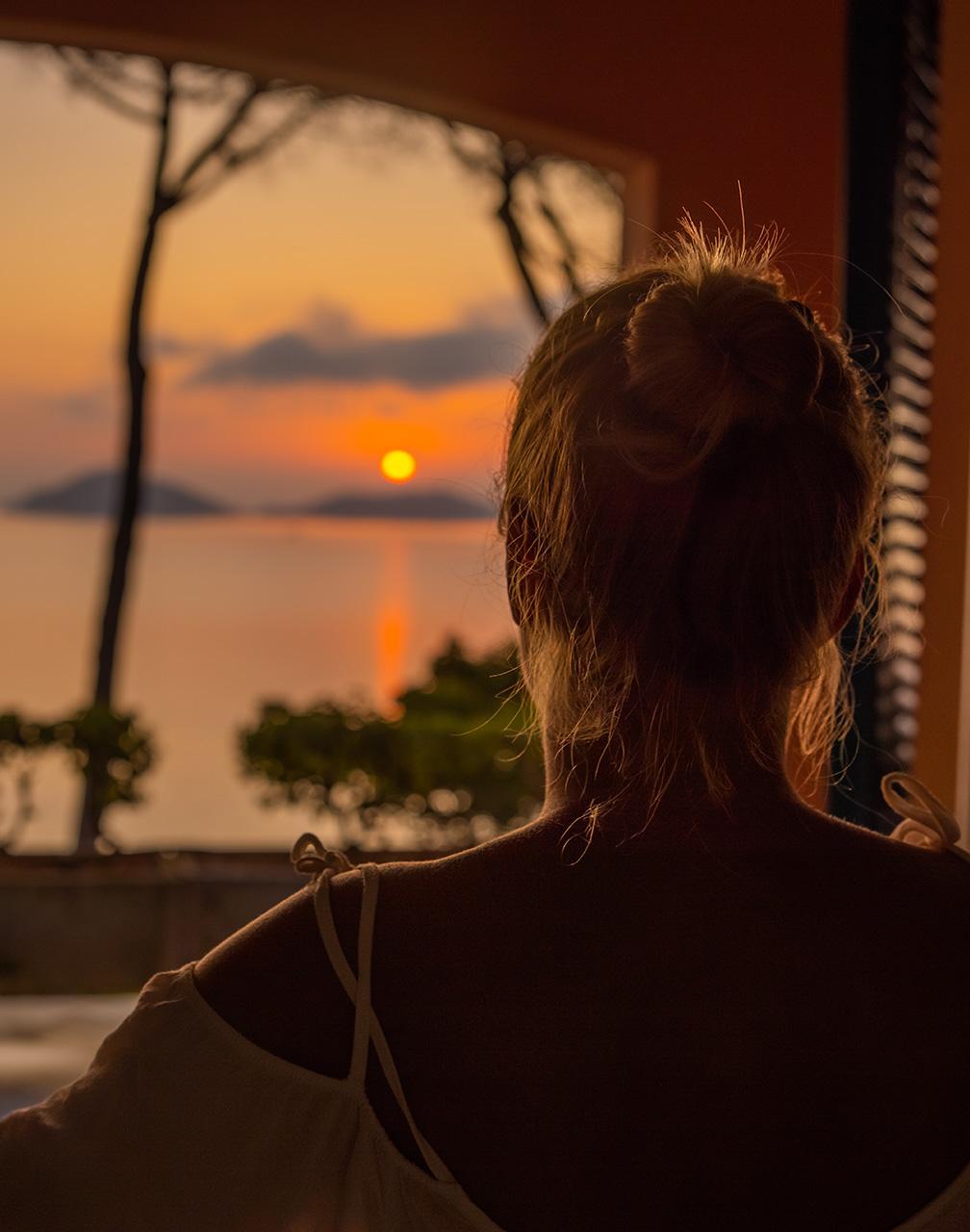 The back of a woman looking out of a window at the sunset above the mountains and sea