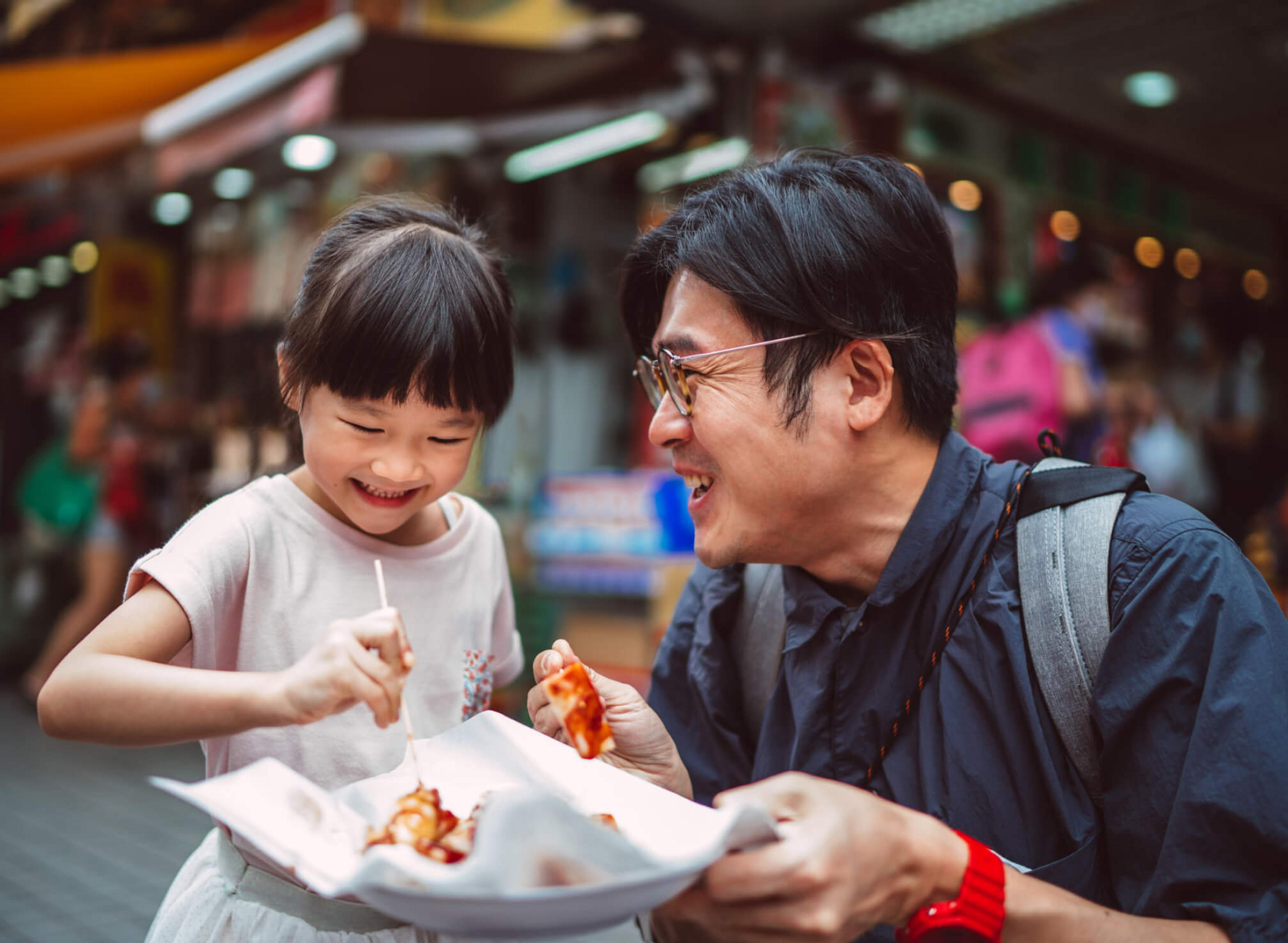 Middle aged man with glasses kneeling next to a young girl smiling and sharing a plate of food in a market