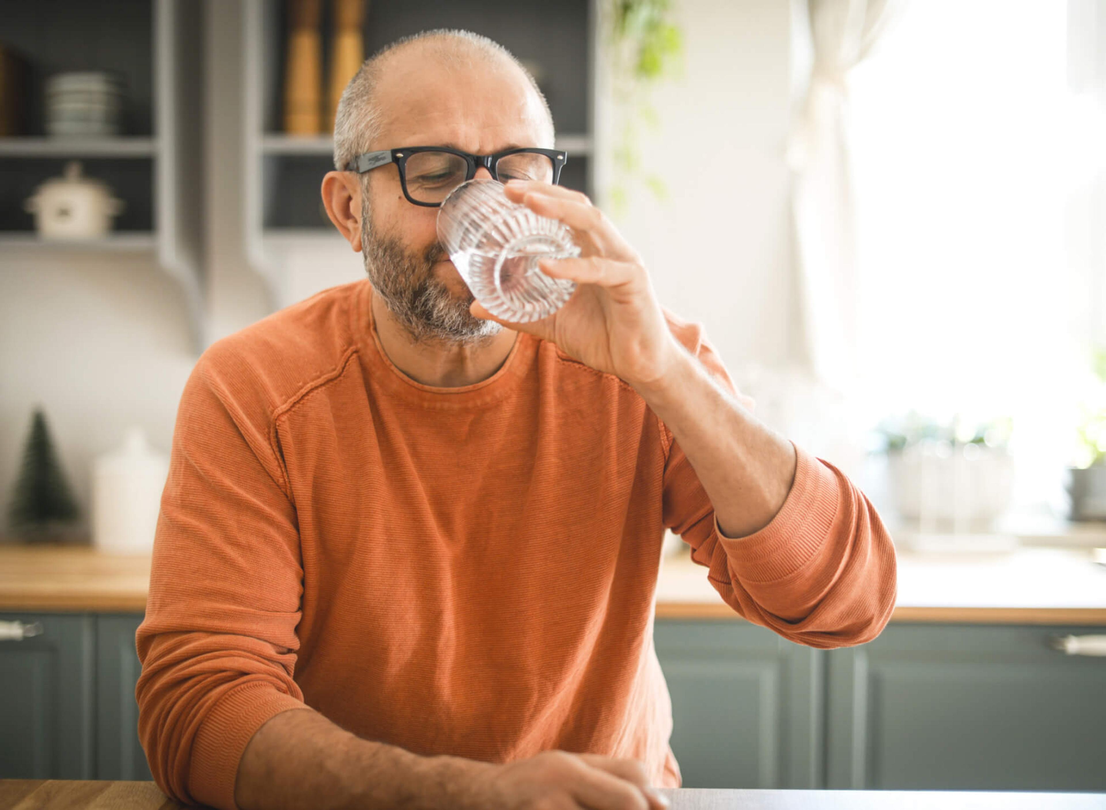 Man sits in a kitchen in an orange jumper drinking a glass of water