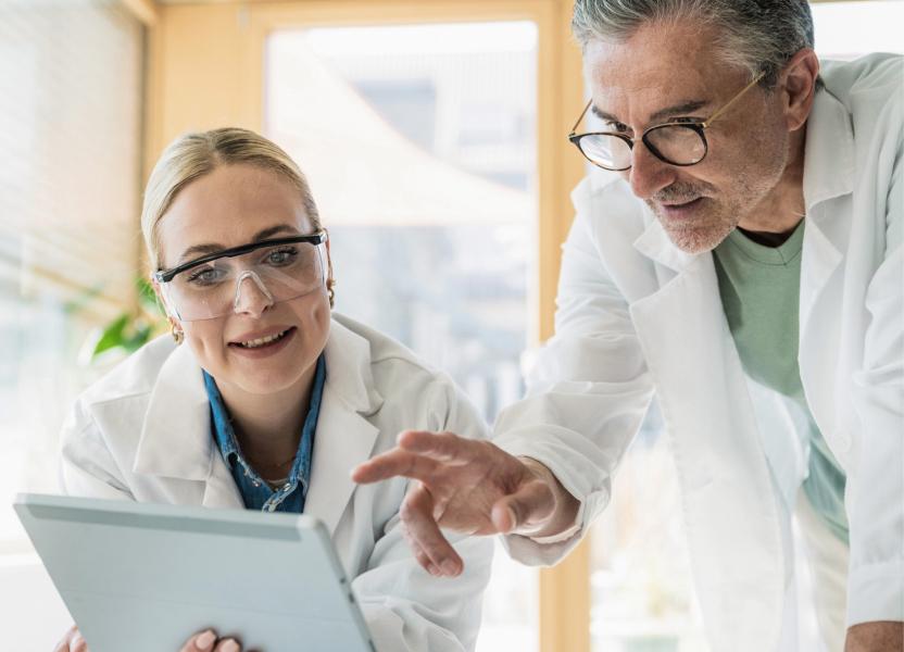 A male healthcare professional in a lab coat points to a tablet held by a female colleague wearing goggles