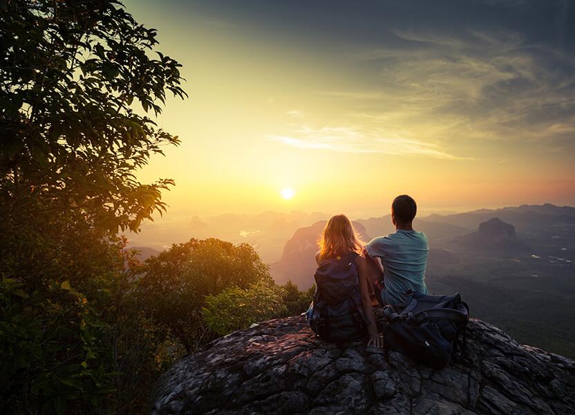 Two people sat on top of a hill looking out towards a mountain range while the sunsets