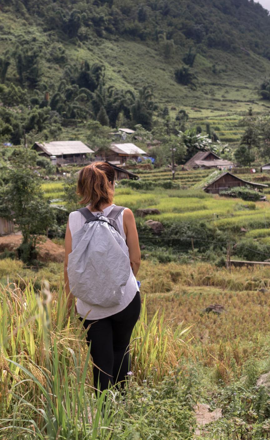Female hiker standing in a field of tall grass with a large backpack looking out to houses at the foot of a mountain