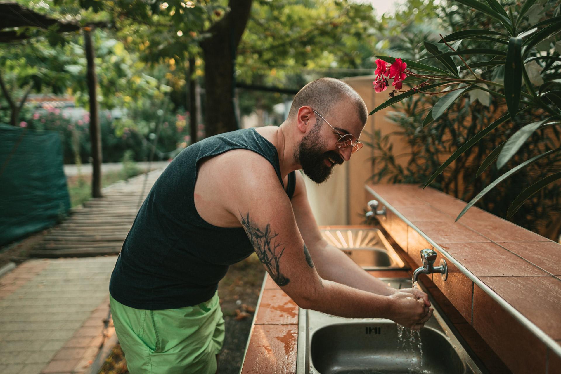 Man with tattoos and glasses smiling and washing his hand in a tropical garden