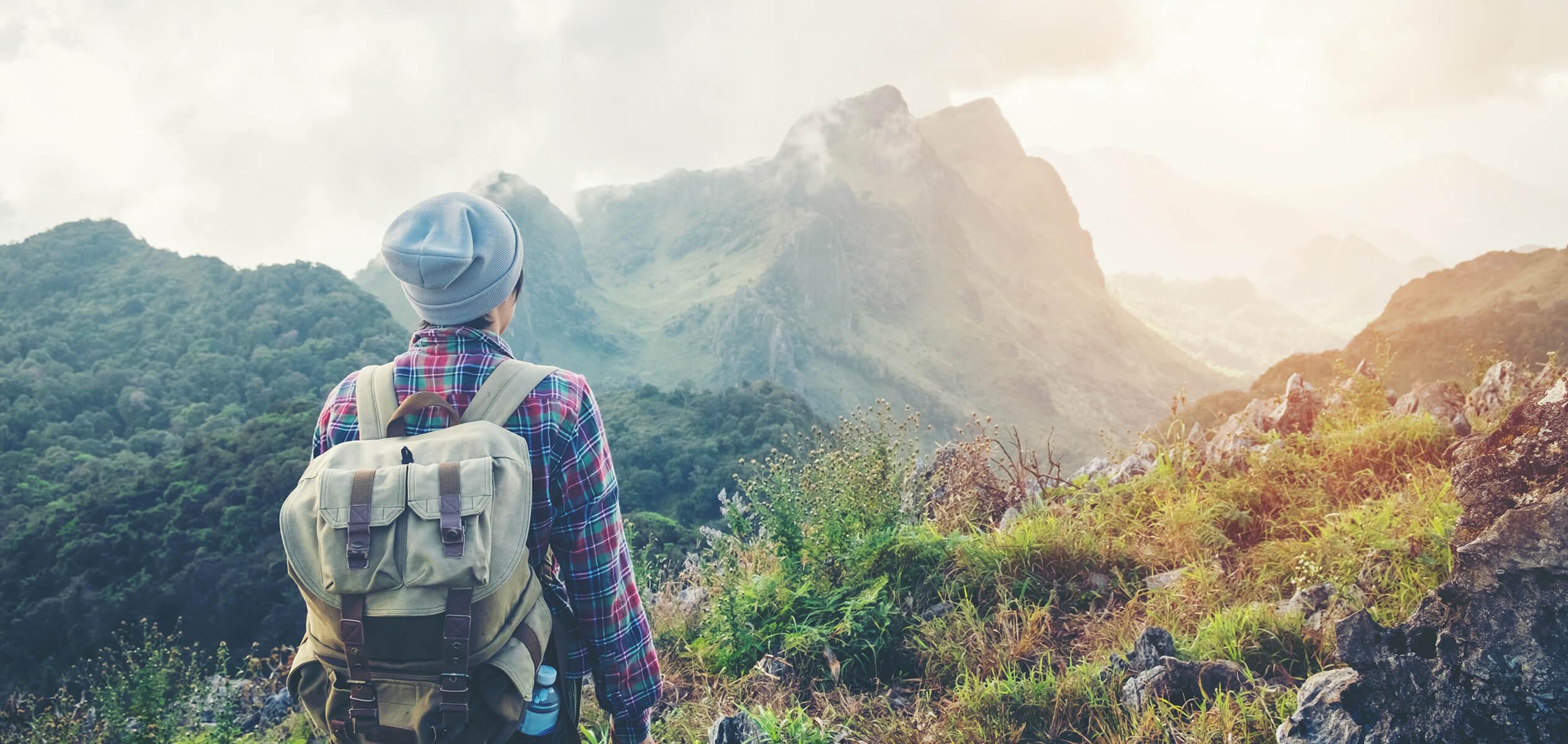 Hiker standing with their back to the camera looking towards a mountain range wearing a backpack