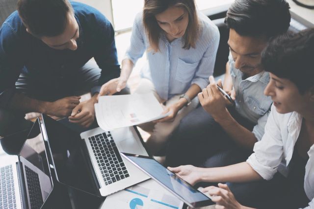 Group of four colleagues sitting together in an office, collaborating over laptops, documents, and a tablet during a team meeting.