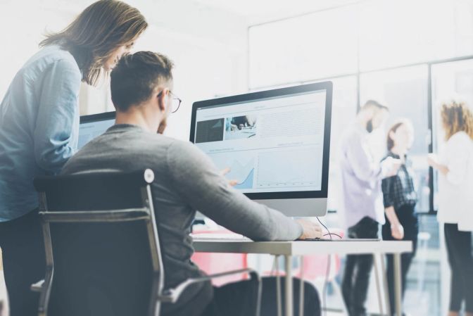 Two coworkers looking at data and charts on a large desktop monitor in a bright modern office, with other colleagues talking in the background.