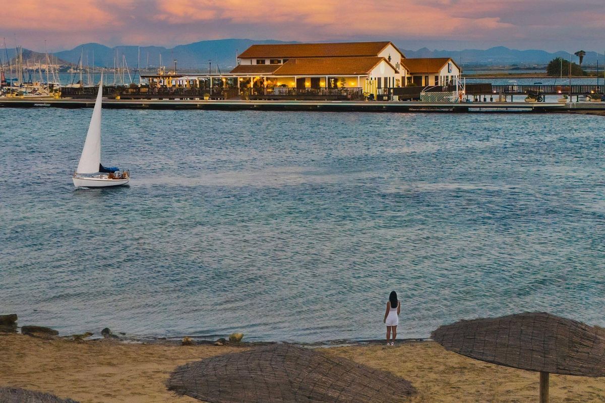 Schilderachtig uitzicht op zonsondergang bij het strand in Los Alcazares, met een zeilboot en een restaurant op de achtergrond.