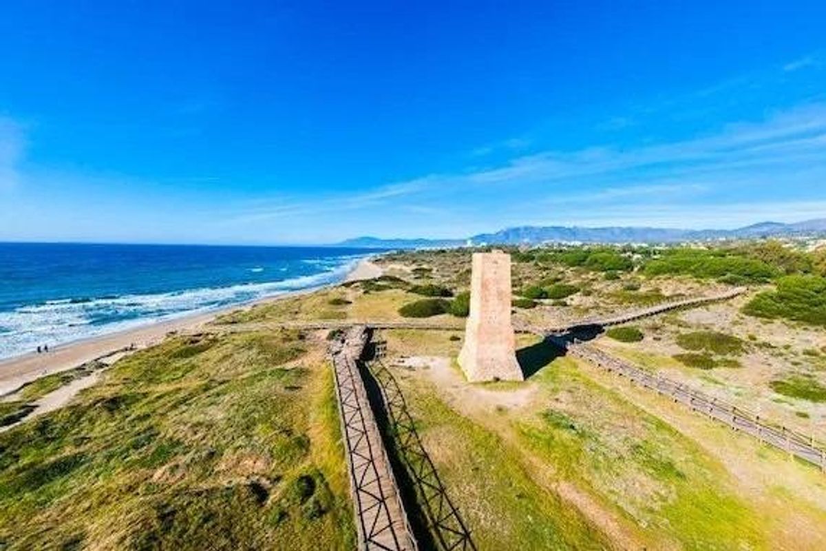Luchtfoto van de historische toren bij het strand in Marbella, Costa del Sol, met golven en bergen op de achtergrond.