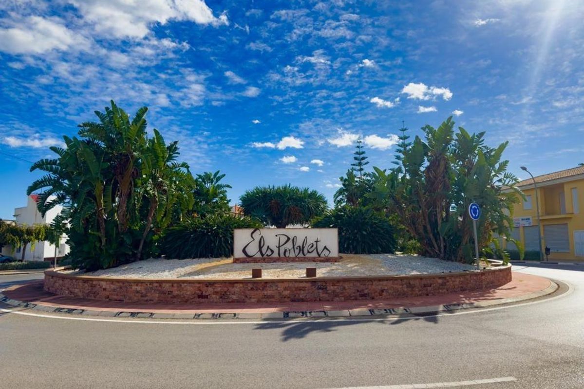 Welkomstbord van Els Poblets, Costa Blanca Noord, omgeven door weelderige palm bomen en blauwe lucht.