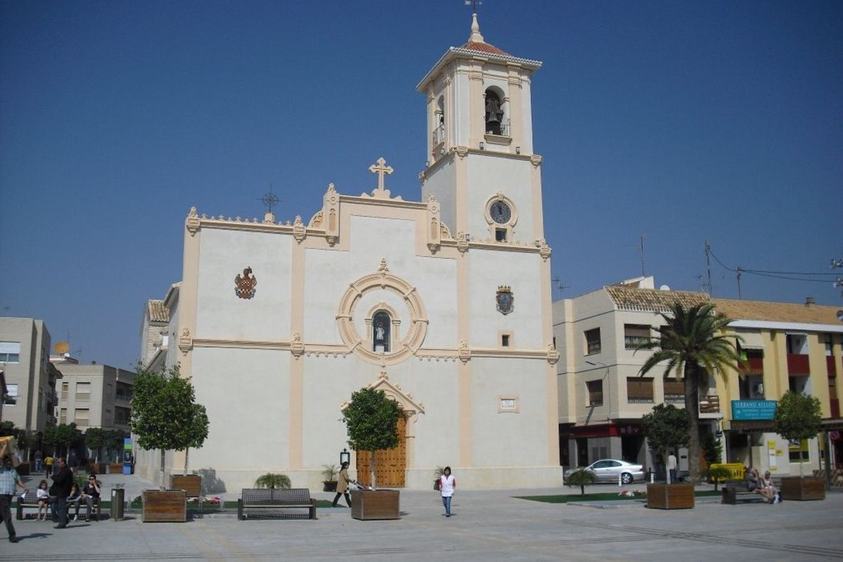 Gevel van een kerkgebouw in san javier, spanje. Stadsplein voor de structuur met bomen, banken en voetgangers.