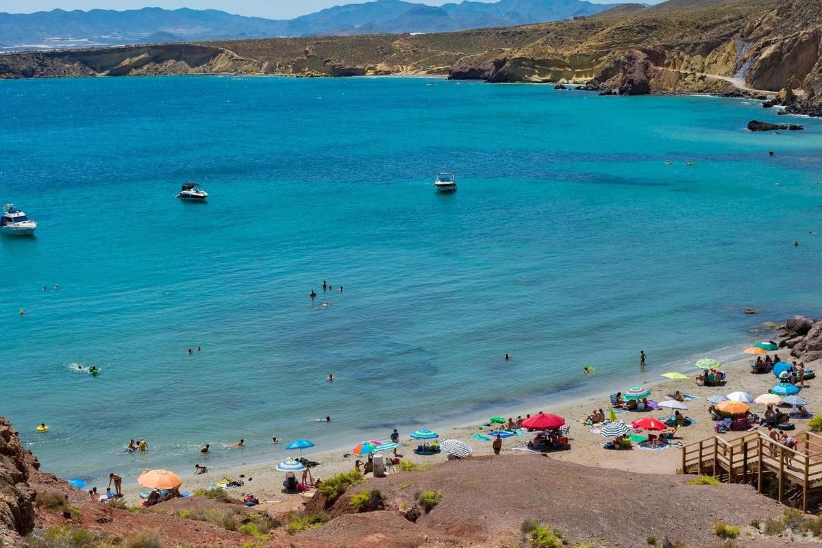 Druk strand in Fuente Álamo met zonaanbidders, kleurrijke parasols en helderblauw water.