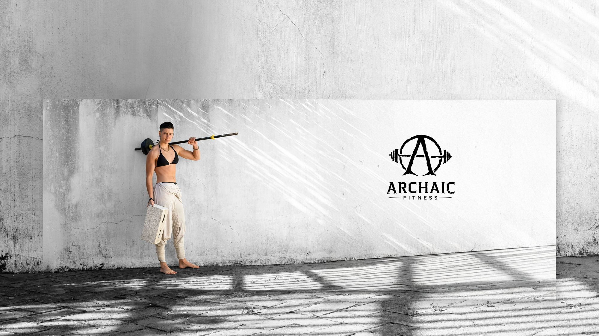 Rebekah-Breeze holding a barbell across the shoulders in front of a concrete wall beside the Archaic Fitness logo