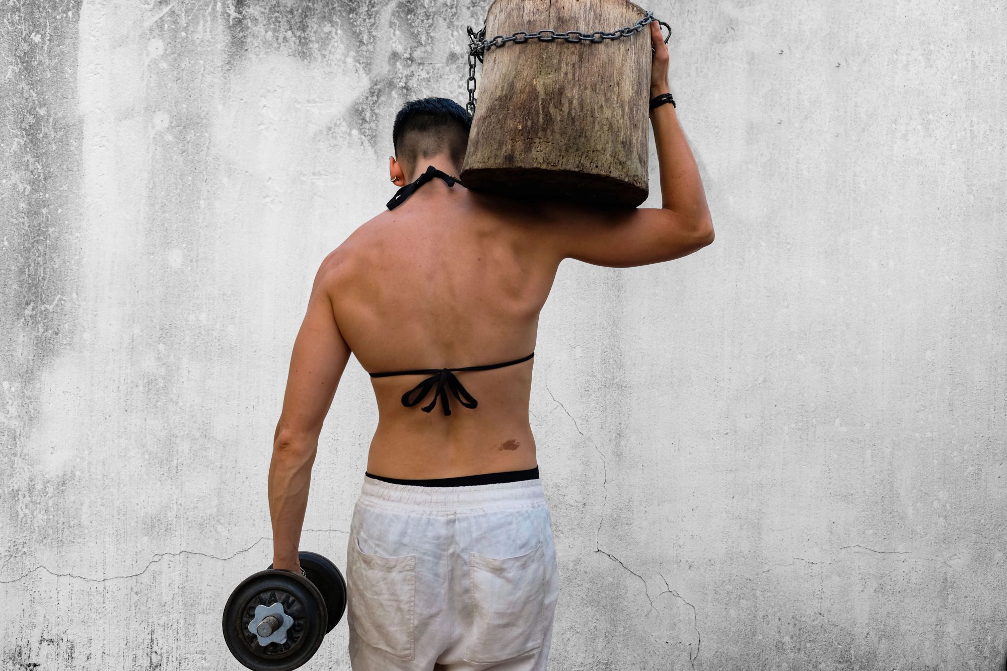 Athlete seen from behind carrying a chained wooden block on one shoulder and a dumbbell in the other hand
