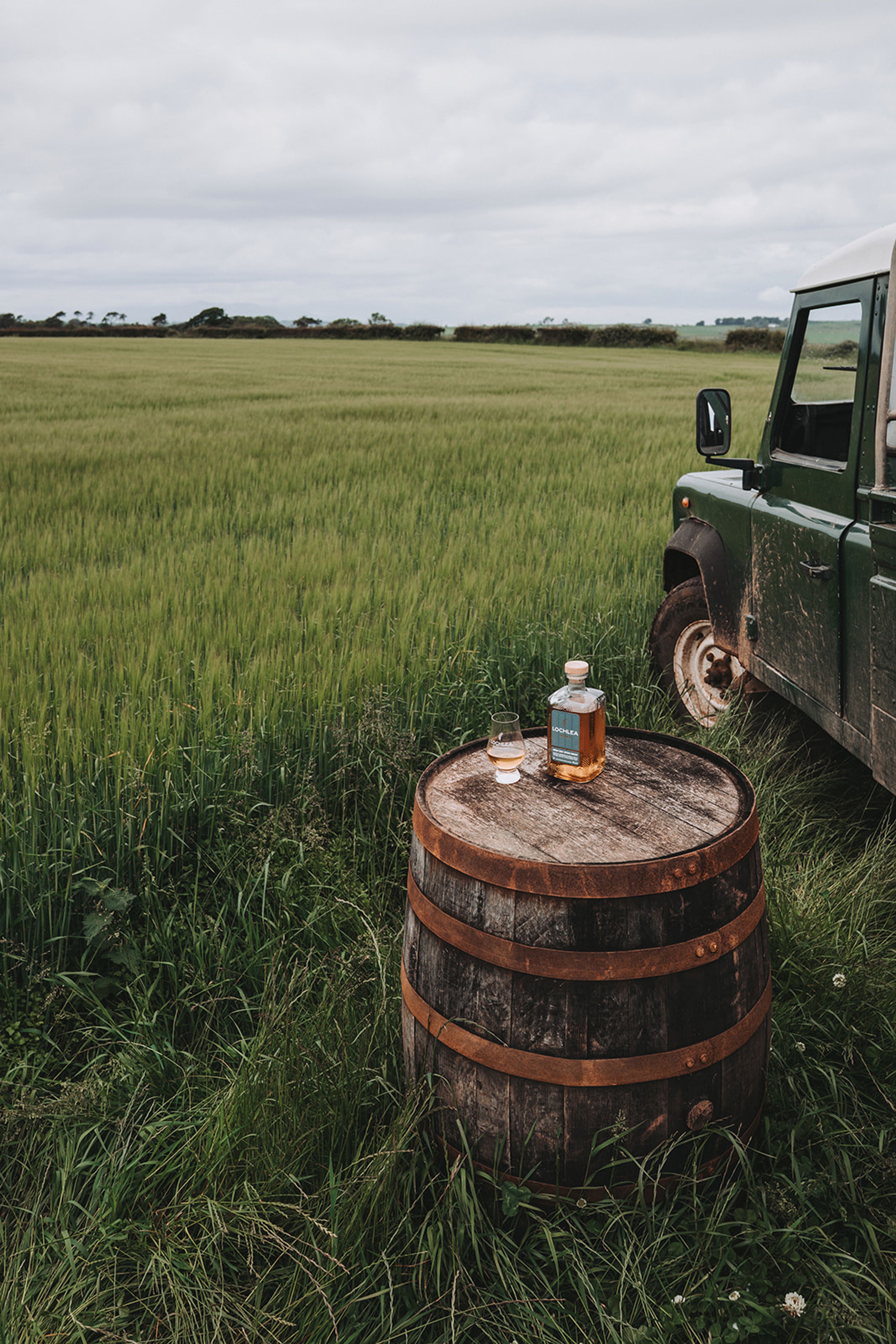 Lochlea whisky bottle and dram on an oak cask beside a Land Rover at the edge of a barley field