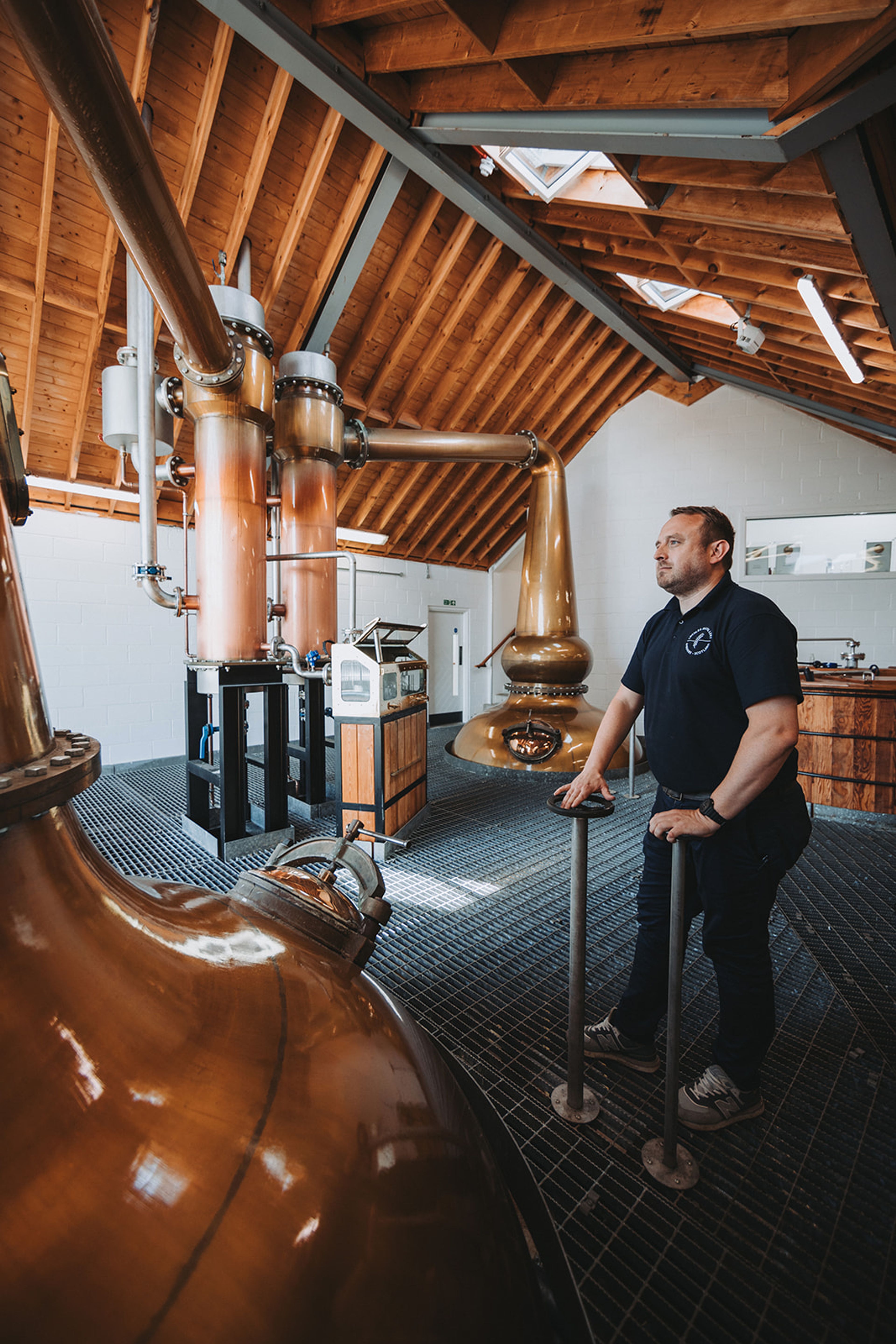 Distiller standing beside copper pot stills in a timber-roofed whisky still house