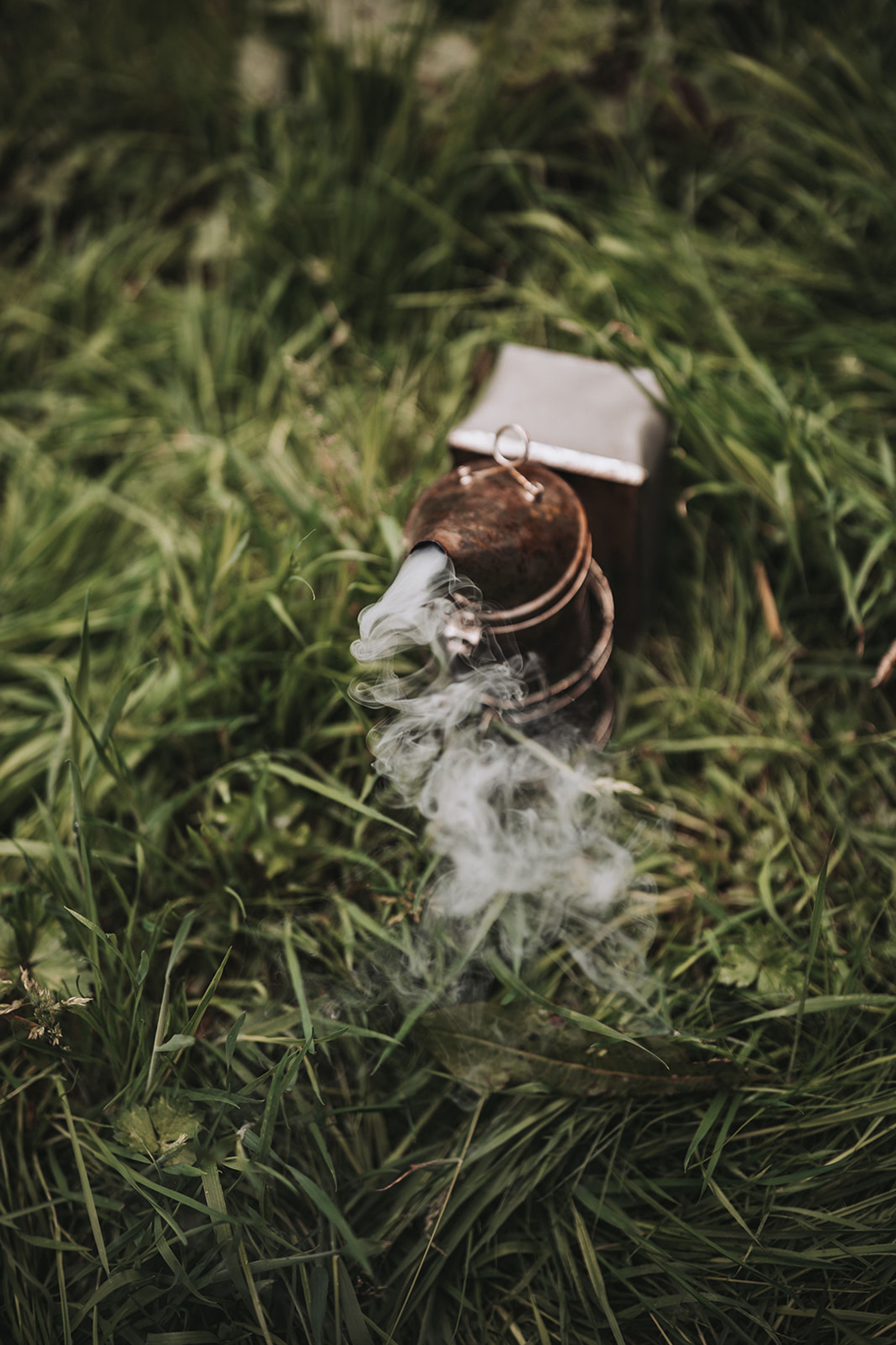 Smoke curling from a rusted lantern lying in long grass