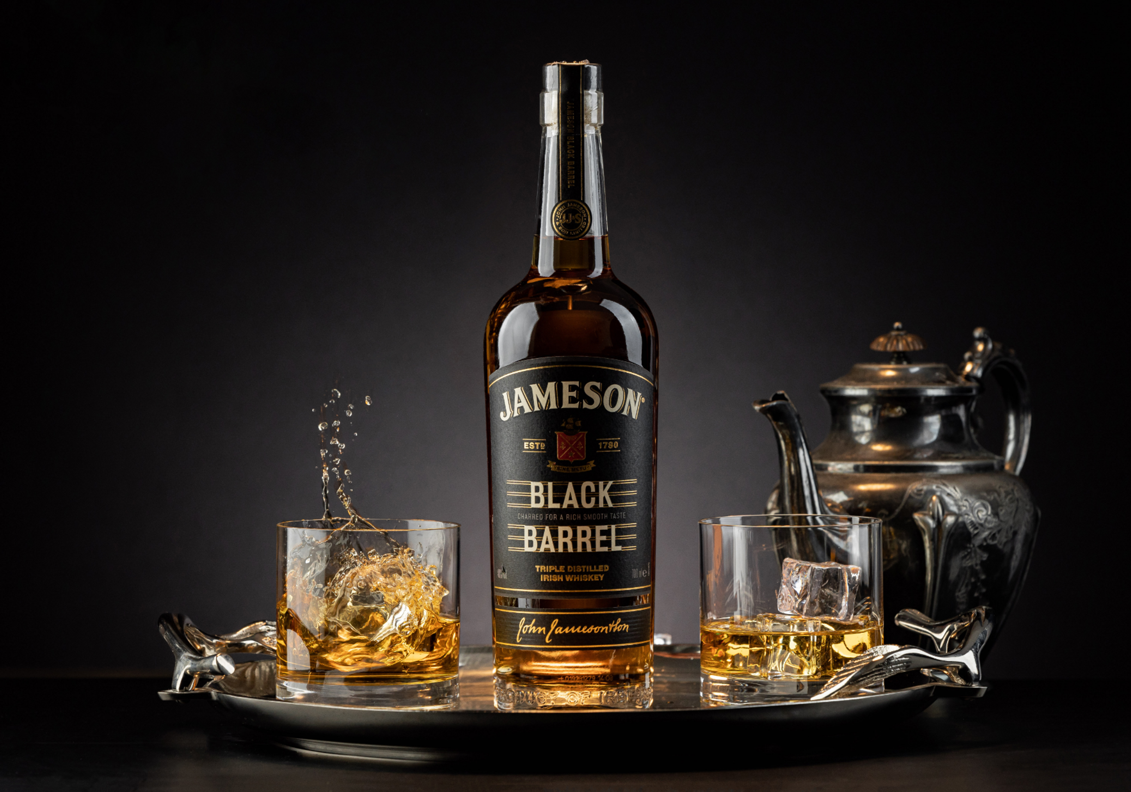 Jameson Black Barrel whiskey bottle with splashing glasses and a silver teapot on a dark set