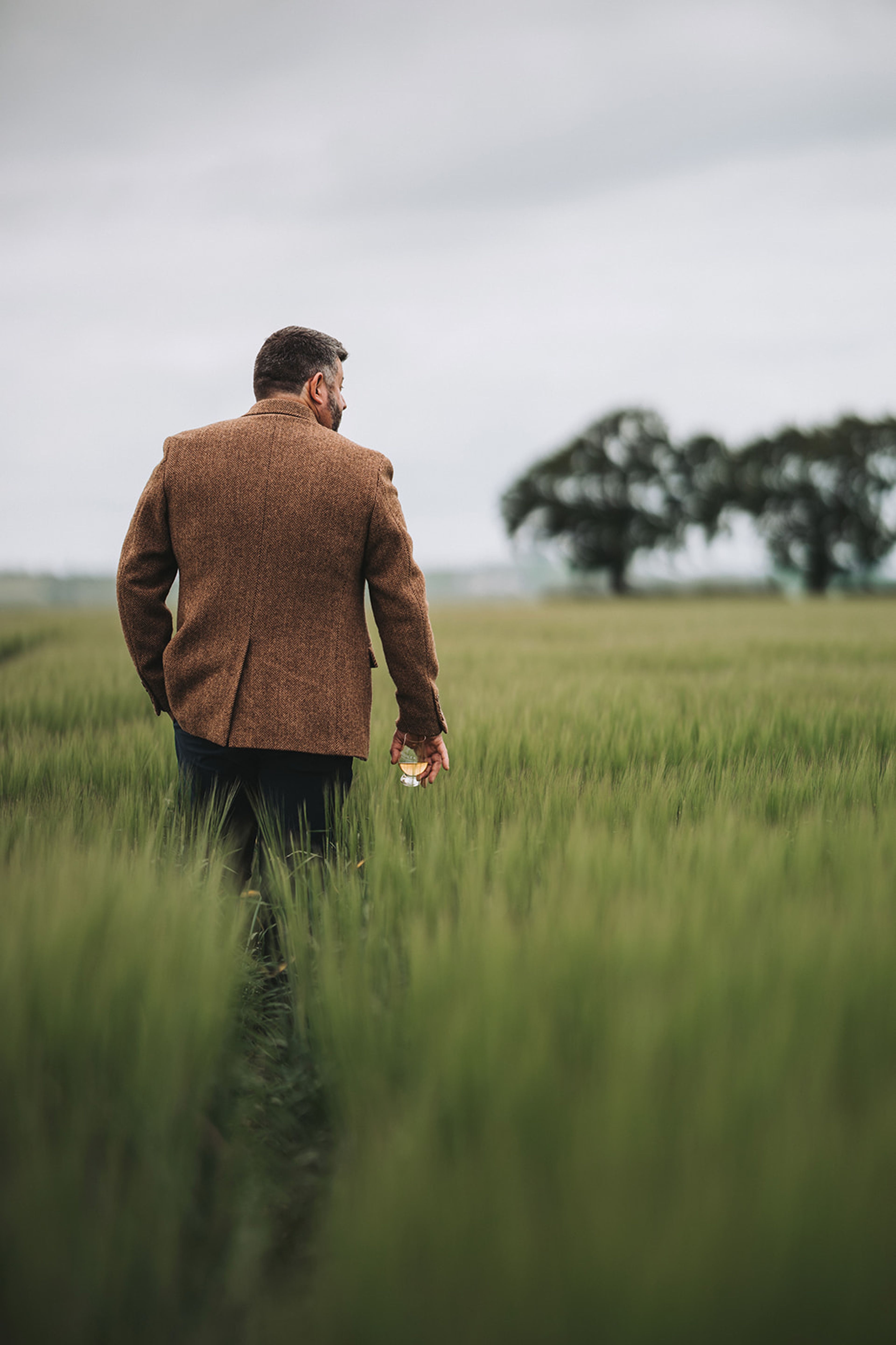 Man in a tweed jacket carrying a whisky glass through a barley field
